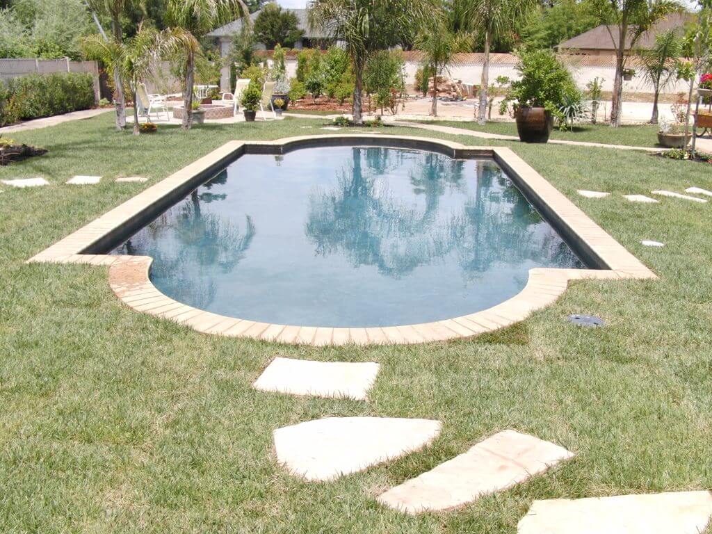 Grecian shaped pool approximately 400 square feet with brick coping surrounded by grass and a stone walkway and palm trees.