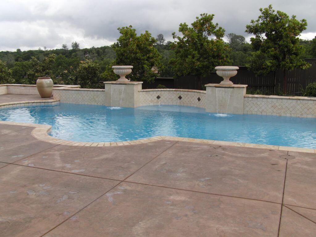 Custom shaped pool with travertine brick coping and a large raised wall with columns, bowls and sheer descent water features.