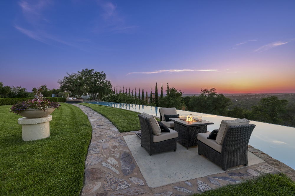Long curved infinity edge pool with a flagstone walkway, fire table and chairs at sunset.