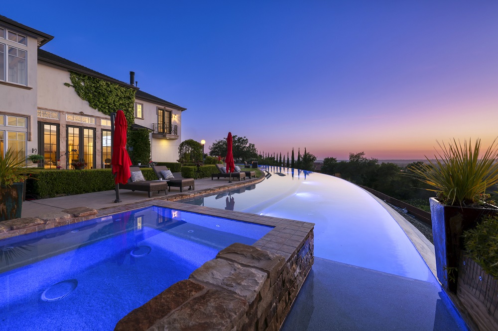 Long curved infinity edge pool with raised spa and long Cabo shelf at sunset.