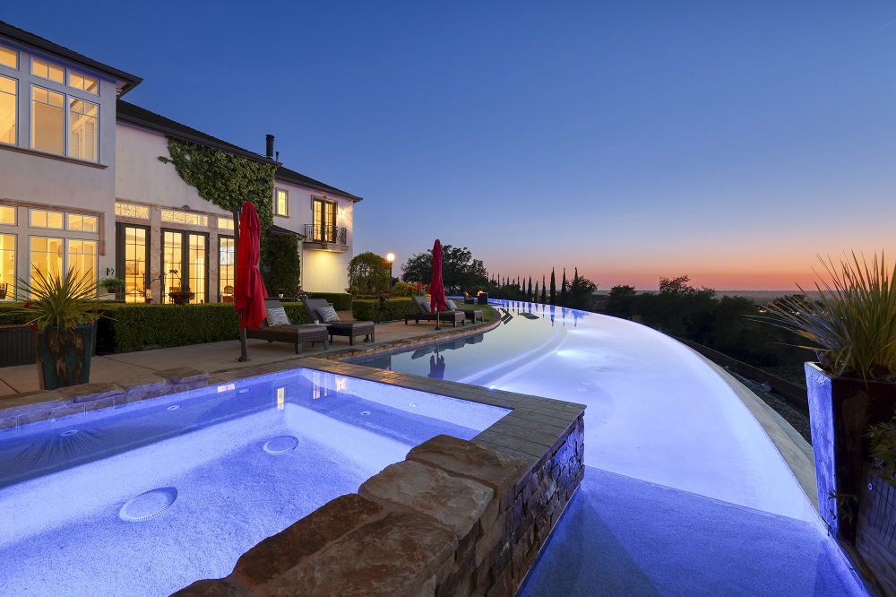 Raised spa with stone that is connected to a long curved infinity edge pool at sunset.