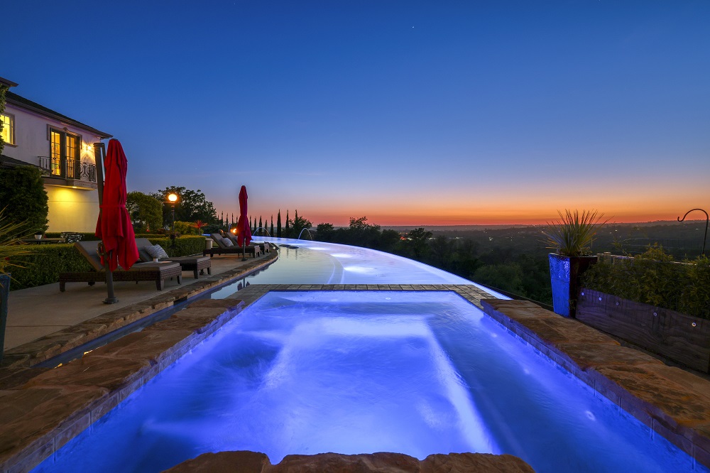 Raised spa is connected to a long curved infinity edge pool at sunset.