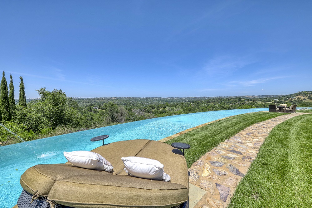 Large curved infinity edge pool with laminars and a grassy area with flagstone walkway and a double person chaise lounge.