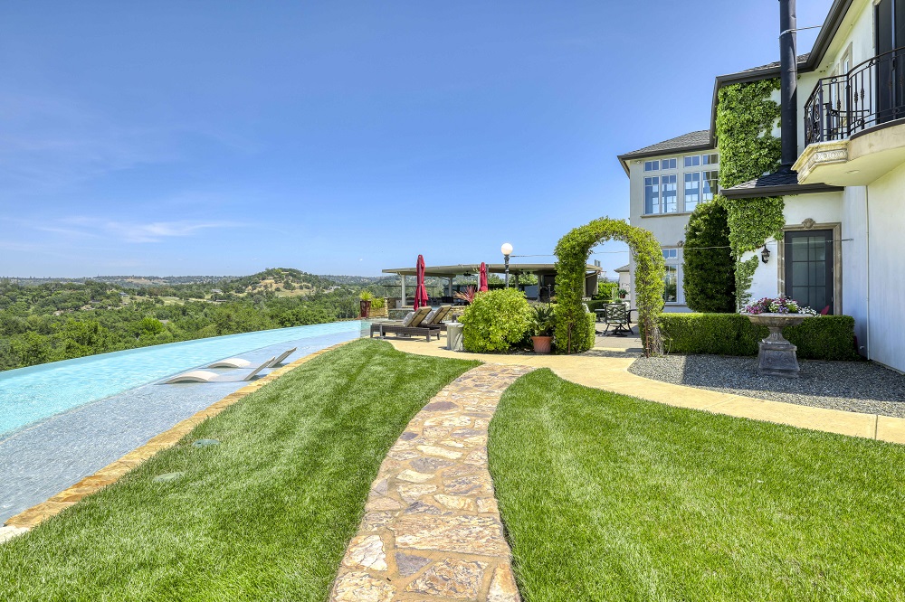 Large curved infinity edge pool with a Cabo shelf with Lounge Ledge chaises next to a grassy area with flagstone walkway.