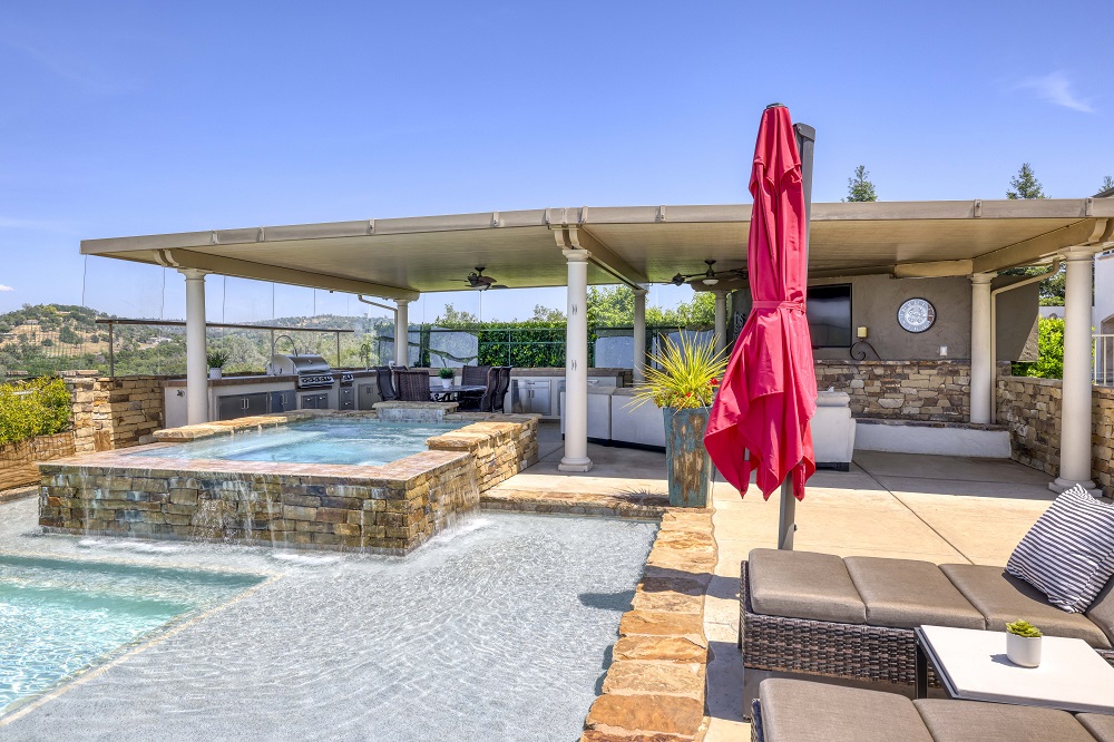 Raised spa with ledgerstone that is spilling over into to a pool with a large Cabo shelf next to a built-in covered kitchen area.