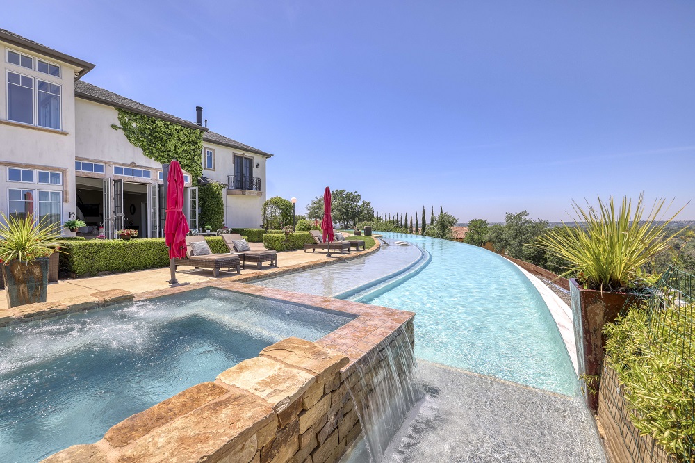 Raised spa with ledgerstone that is spilling over into to a long curved infinity edge pool during the day.