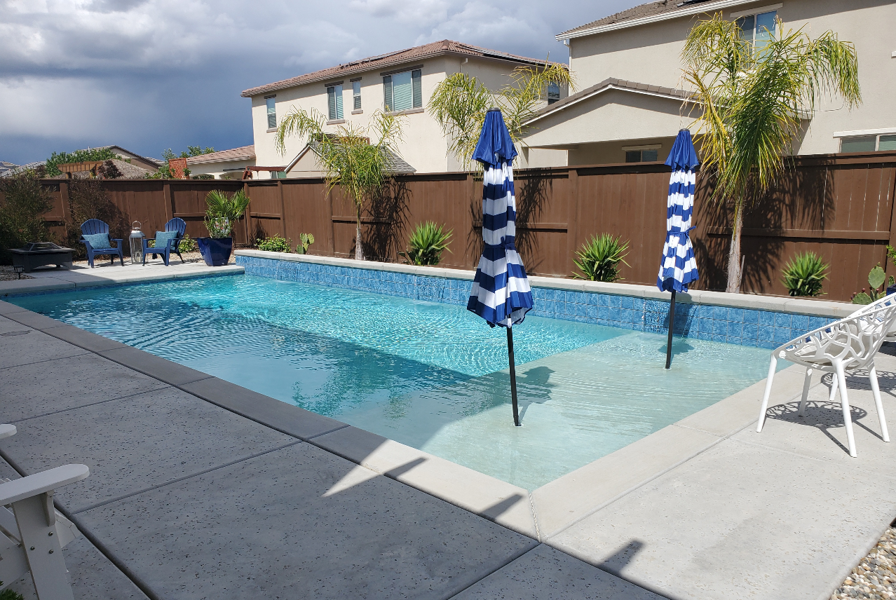 Rectangular shaped pool with raised wall with sheer descent water features and large Cabo shelf with two umbrellas.