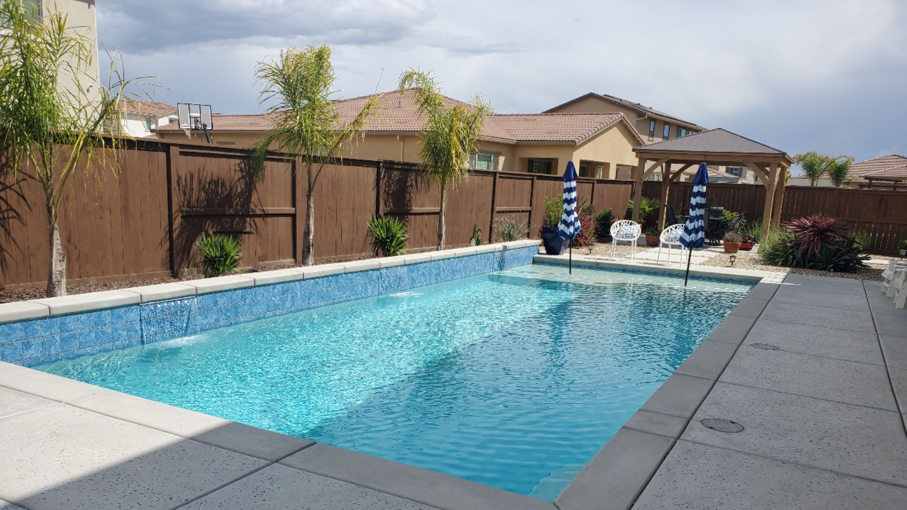 Rectangular shaped pool with a blue tile faced raised wall with three sheer descent water features and a large Cabo shelf with two umbrellas plus a seating area with loggia.