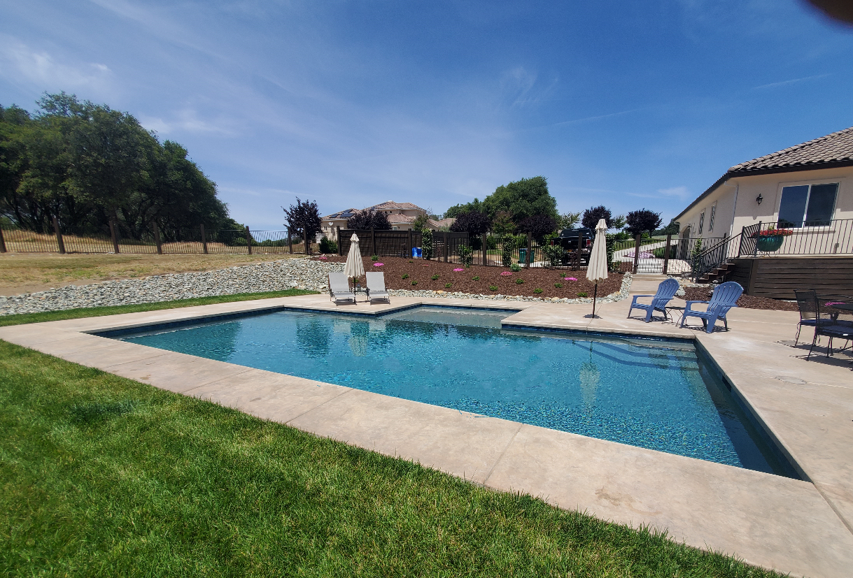 Rectangular shaped pool with larged bumped out Cabo shelf wtih concrete decking and grass next to a house on a sloped lot.