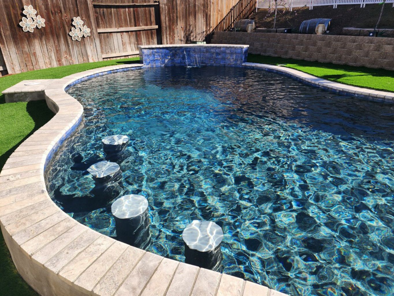 Partially raised freeform shaped pool with raised wall with sheer descent water feature, four barstools and travertine stone coping.