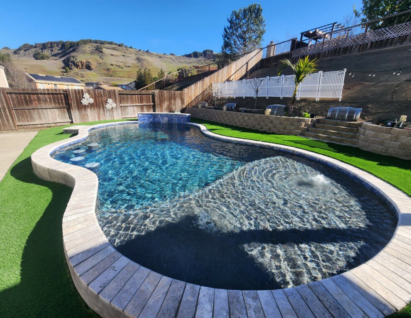Partially raised freeform shaped pool with raised wall with sheer descent water feature, four barstools, large Cabo shelf and travertine stone coping.