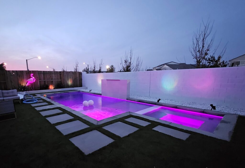 Rectangular pool with spa, raised wall with sheer descent, and colored LED lighting at night